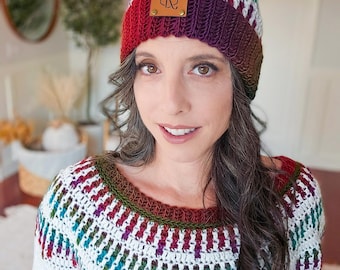 May include: A white crocheted hat with a burgundy pom pom and a brown leather patch with the initials "CR" on it. The hat is worn by a person with long dark hair and a white crocheted sweater with a colorful pattern.
