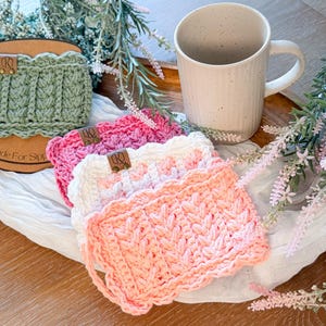 May include: A collection of handmade crochet mug rugs in shades of pink, white, and green, with a textured pattern and scalloped edges. Each rug features a small leather tag. A beige mug sits nearby. The text "Made For Sips" is visible.