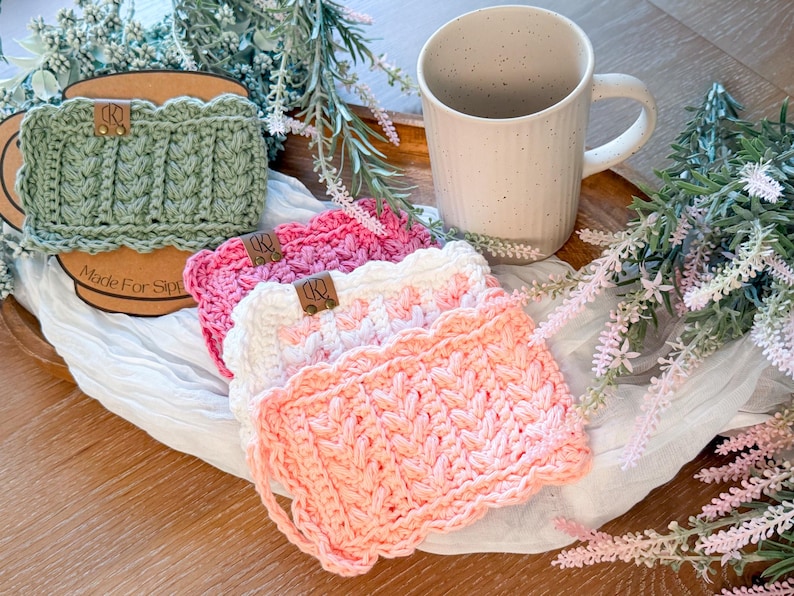 May include: A collection of handmade crochet mug rugs in shades of pink, white, and green, with a textured pattern and scalloped edges. Each rug features a small leather tag. A beige mug sits nearby. The text "Made For Sips" is visible.