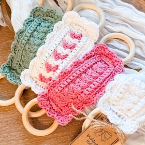 May include: A collection of handmade crochet dish towel holders in various colors, including green, white, and pink. Each holder features a wooden ring and a decorative crochet design. A small tag reads "Dish Towel Holder".