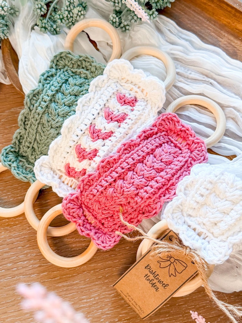 May include: A collection of handmade crochet dish towel holders in various colors, including green, white, and pink. Each holder features a wooden ring and a decorative crochet design. A small tag reads "Dish Towel Holder".