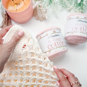 Hands crocheting with ivory yarn, two skeins of Cottage Cotton yarn nearby, a lit candle, and a leafy plant in the background—perfect inspiration for a handmade gift, all set on a white surface.