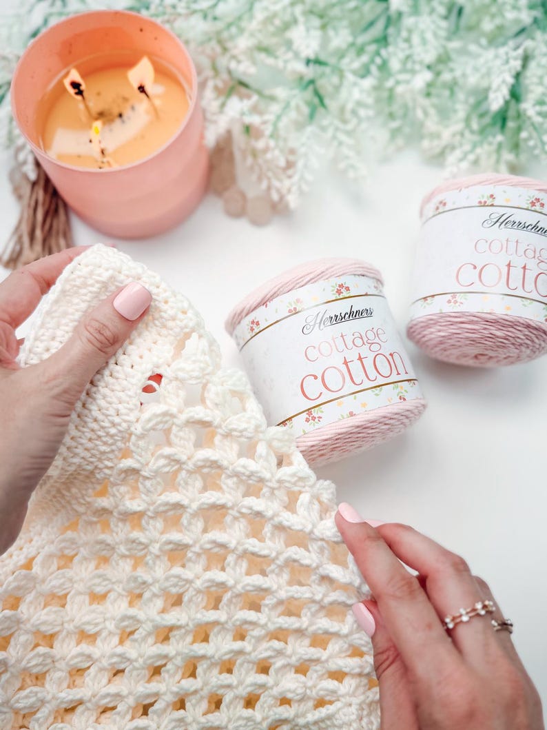 Hands crocheting with ivory yarn, two skeins of Cottage Cotton yarn nearby, a lit candle, and a leafy plant in the background—perfect inspiration for a handmade gift, all set on a white surface.