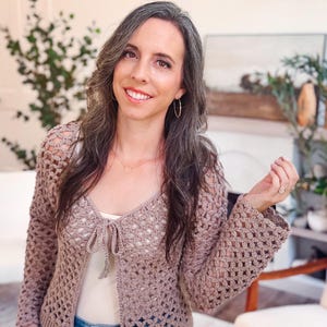 May include: A light brown, long-sleeved crochet cardigan featuring an open-knit, repeating floral pattern throughout the body and sleeves. The garment includes a front tie closure at the neckline and a solid, ribbed knit border along the bottom hem.