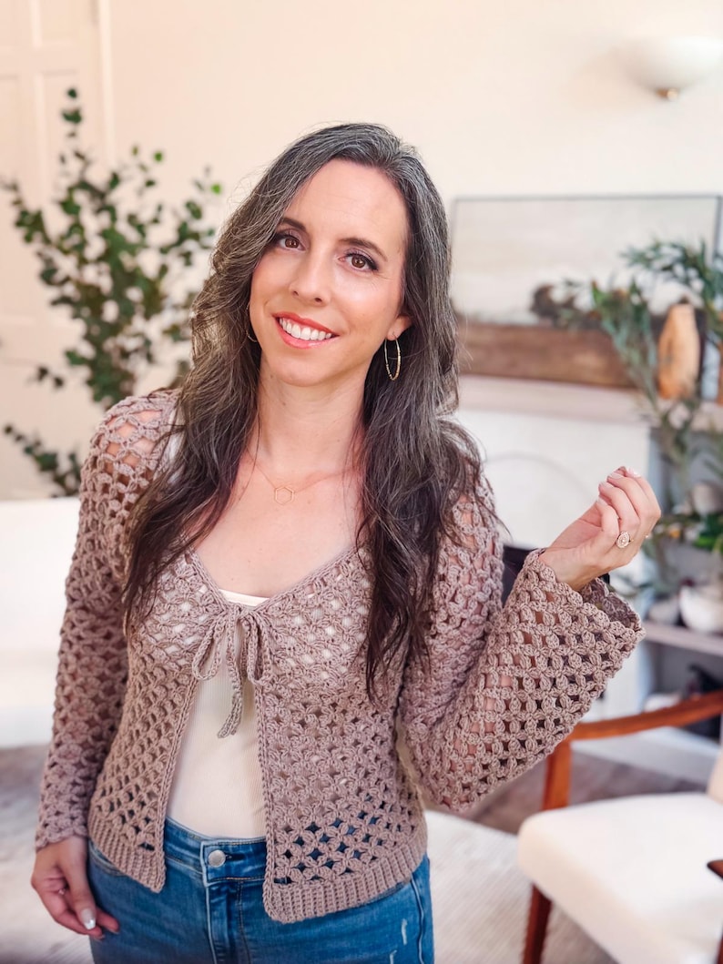 May include: A light brown, long-sleeved crochet cardigan featuring an open-knit, repeating floral pattern throughout the body and sleeves. The garment includes a front tie closure at the neckline and a solid, ribbed knit border along the bottom hem.