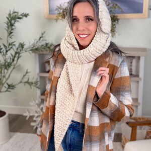 May include: A woman wearing a cream-coloured knitted hood and scarf set. She is also wearing a white sweater, blue jeans, and a brown, orange, and white plaid coat. The background includes a painting, a plant, and a fireplace.