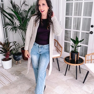 A person with long hair is standing indoors near plants, donning unique jewellery that complements their long beige cardigan. They sport a dark top, light blue jeans, and black shoes. The scene features a tiled floor and glass doors in the background.
