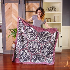 Op de afbeelding: Een groot, handgebreid deken met een geometrisch patroon in tinten roze, wit en grijs. Het deken heeft een herhalend zeshoekig ontwerp met ingewikkelde details. Het deken wordt vastgehouden door een persoon in een grijs shirt.