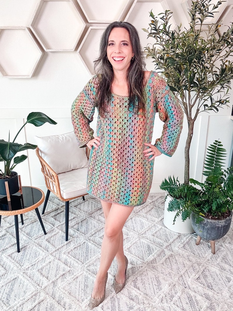 A person wearing a colorful granny crochet dress and unique jewellery stands indoors near a chair and various plants, smiling at the camera.