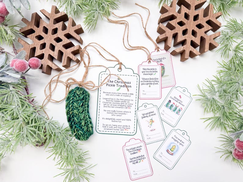 May include: A green knitted pickle ornament with a brown twine string, accompanied by several white paper gift tags featuring illustrations of pickles and text explaining The Christmas Pickle Tradition, including spaces for to and from names.