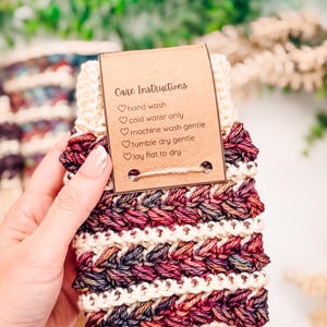 May include: A hand-knitted item in burgundy, cream, and dark blue. It features a textured, braided pattern and a tag with care instructions: hand wash, cold water only, machine wash gentle, tumble dry gentle, and lay flat to dry.