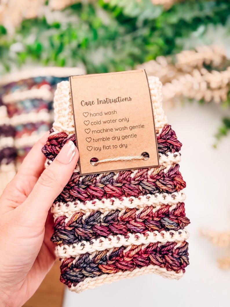 May include: A hand-knitted item in burgundy, cream, and dark blue. It features a textured, braided pattern and a tag with care instructions: hand wash, cold water only, machine wash gentle, tumble dry gentle, and lay flat to dry.