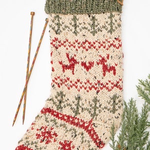 Op de afbeelding: Een gebreide kerstsok met een groen, rood en wit patroon met rendieren. De sok heeft een bruine leren lus aan de bovenkant en wordt vergezeld door twee breinaalden.