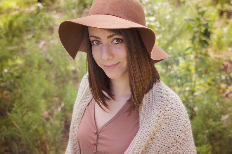 A woman wearing a cream crochet shawl wrap and hat, in a field of greenery.