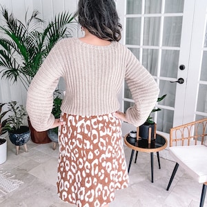 A person standing in a room with hands on hips, facing away from the camera, wearing a beige crochet sweater and a leopard print skirt paired with unique jewellery.