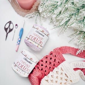 Puede incluir: Dos ovillos de hilo Herrschners cottage cotton en rosa claro y blanco, un ganchillo azul con lunares rosas, un marcador de puntos peque&ntilde;o, tijeras de metal oscuro y un bolso parcialmente tejido a ganchillo en hilo de algod&oacute;n rosa y blanco.