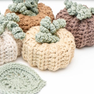 Four ribbed crochet pumpkins in cream, beige, tan and taupe with curly green stems beside a sage leaf coaster