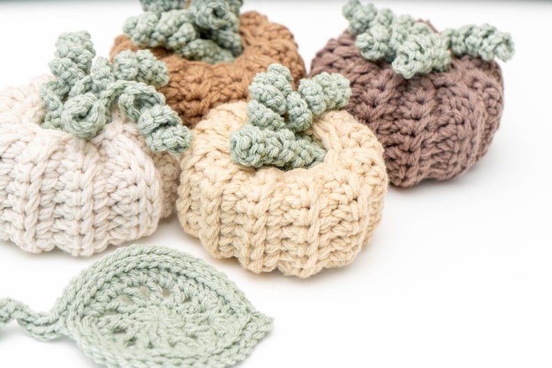 Four ribbed crochet pumpkins in cream, beige, tan and taupe with curly green stems beside a sage leaf coaster