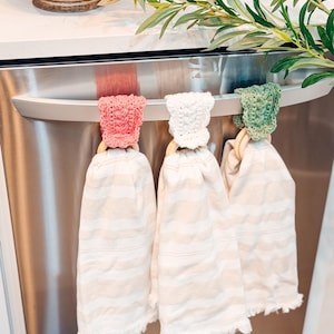 May include: Three kitchen towels hanging on a stainless steel appliance handle. Each towel is cream-colored with horizontal stripes and a fringed edge. The towels are held by wooden rings and topped with crocheted caps in pink, white, and green.
