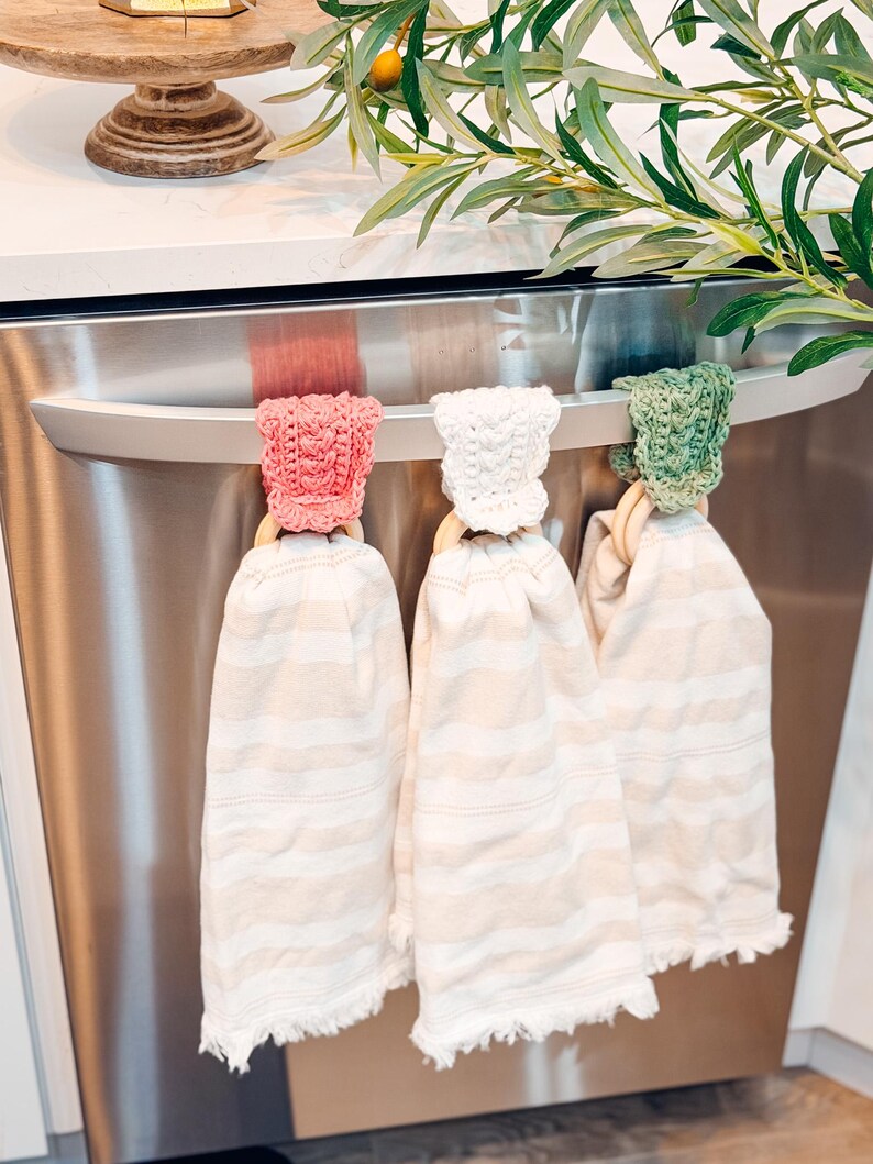 May include: Three kitchen towels hanging on a stainless steel appliance handle. Each towel is cream-colored with horizontal stripes and a fringed edge. The towels are held by wooden rings and topped with crocheted caps in pink, white, and green.