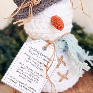 Close-up of a white crochet snowman in a gray hat and blue scarf wearing a funny emotional support snowman tag