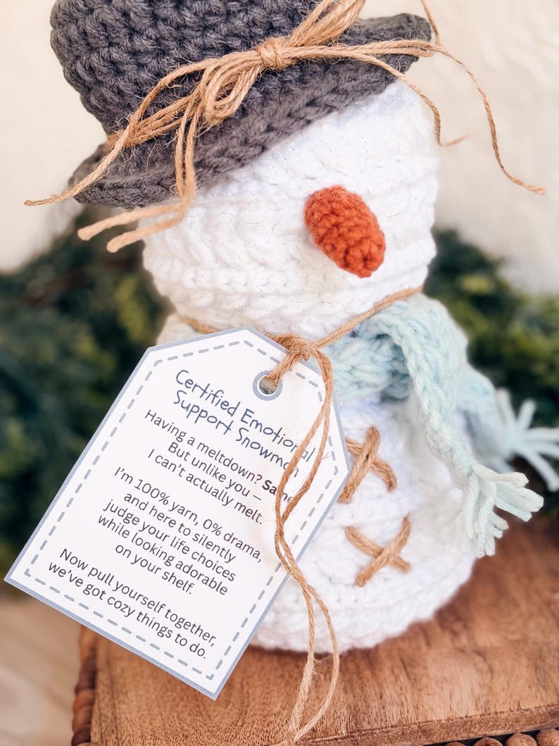 Close-up of a white crochet snowman in a gray hat and blue scarf wearing a funny emotional support snowman tag