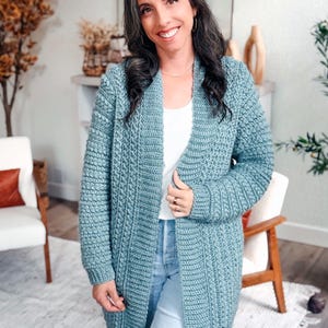 May include: A light blue, open-front crochet cardigan. The long-sleeved sweater has a textured stitch pattern and is worn over a white top and blue jeans. The cardigan is knee-length and appears to be handmade.