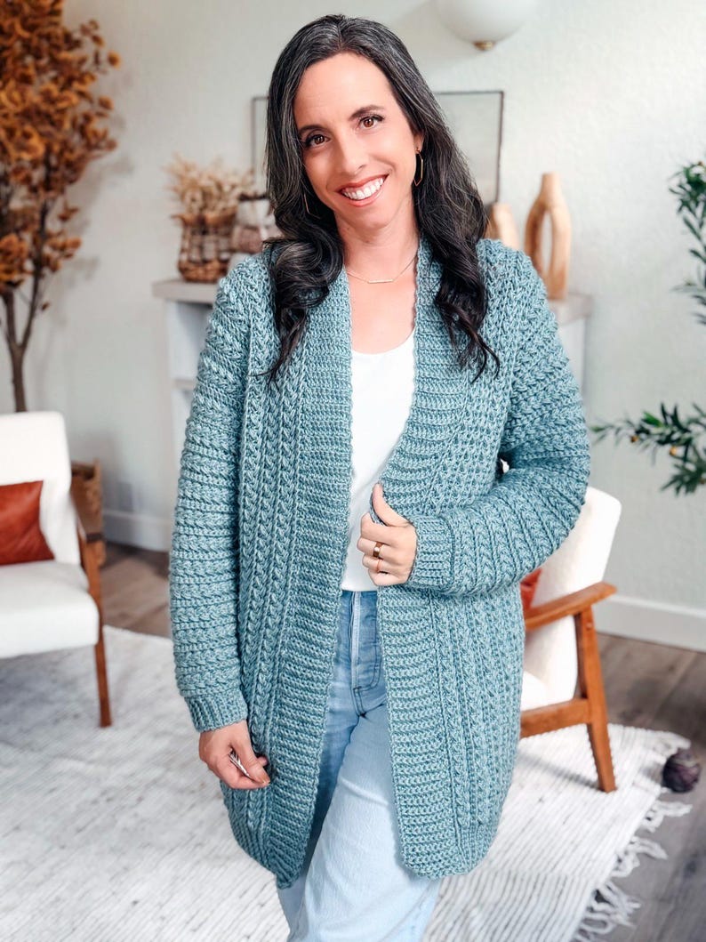 May include: A light blue, open-front crochet cardigan. The long-sleeved sweater has a textured stitch pattern and is worn over a white top and blue jeans. The cardigan is knee-length and appears to be handmade.