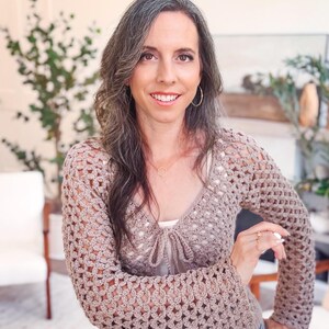 May include: A beige long-sleeved crochet cardigan featuring an openwork floral pattern, a V-neckline, and a front tie closure. The garment is worn over a white top and paired with blue denim jeans, showcasing a textured knit design throughout.