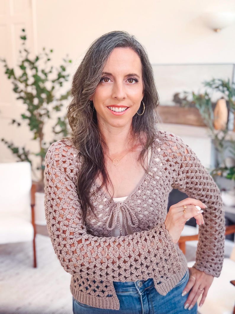 May include: A beige long-sleeved crochet cardigan featuring an openwork floral pattern, a V-neckline, and a front tie closure. The garment is worn over a white top and paired with blue denim jeans, showcasing a textured knit design throughout.