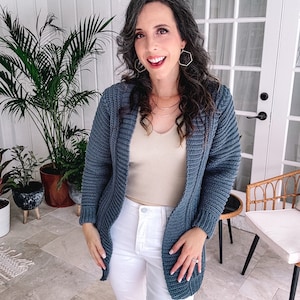 A person with long dark hair wearing a blue crocheted cardigan and white pants stands indoors near plants and furniture, adorned with unique jewellery.