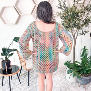 A person with brow and gray hair wearing a colorful crocheted sweater dress stands with their back to the camera, surrounded by plants and hexagonal wall decor, exuding a sense of charm in their vintage clothes.