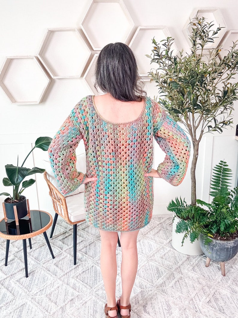 A person with brow and gray hair wearing a colorful crocheted sweater dress stands with their back to the camera, surrounded by plants and hexagonal wall decor, exuding a sense of charm in their vintage clothes.