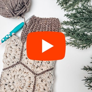 May include: A crochet project featuring a cream-colored hexagonal motif panel with brown yarn borders, a brown ribbed cuff, a ball of brown yarn, and a teal crochet hook with white polka dots, accompanied by green evergreen branches.
