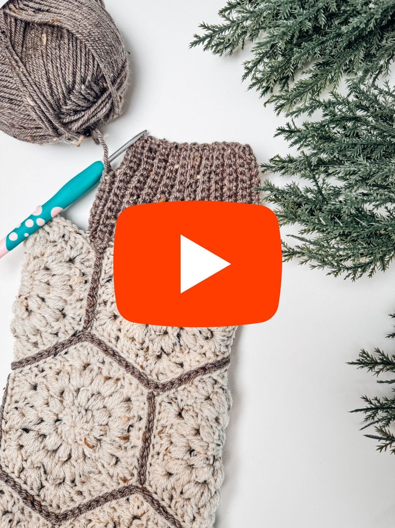 May include: A crochet project featuring a cream-colored hexagonal motif panel with brown yarn borders, a brown ribbed cuff, a ball of brown yarn, and a teal crochet hook with white polka dots, accompanied by green evergreen branches.