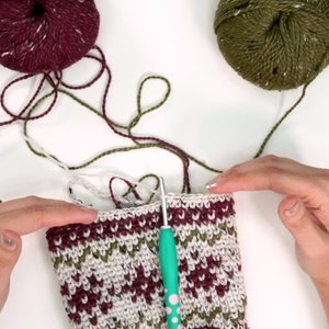 Op de afbeelding: Een gedeeltelijk gehaakt werkstuk met een fair isle-patroon in bordeauxrood, olijfgroen en cr&egrave;mekleurig garen. Een turquoise haaknaald is in de bovenrand gestoken. Twee bollen bijpassend bordeauxrood en olijfgroen garen liggen erboven.