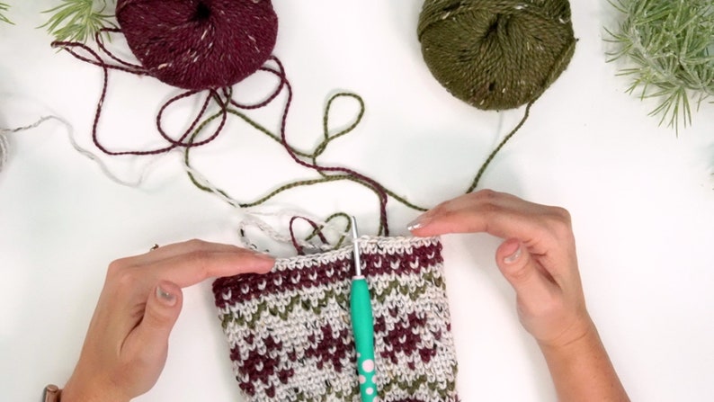 Op de afbeelding: Een gedeeltelijk gehaakt werkstuk met een fair isle-patroon in bordeauxrood, olijfgroen en cr&egrave;mekleurig garen. Een turquoise haaknaald is in de bovenrand gestoken. Twee bollen bijpassend bordeauxrood en olijfgroen garen liggen erboven.