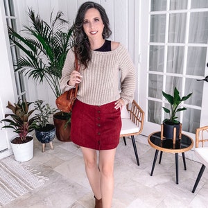 A woman standing indoors, smiling at the camera, dressed in a beige crochet sweater, red skirt, and brown boots, holding a handmade gift, with plants and furniture in the background.