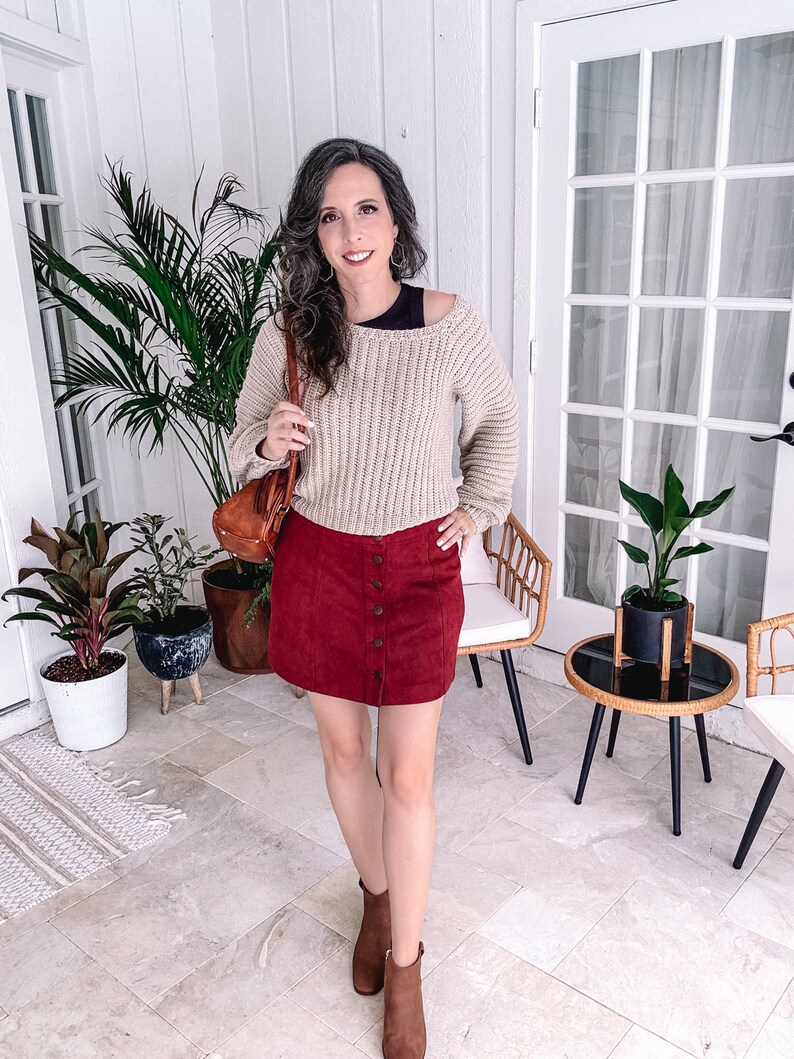 A woman standing indoors, smiling at the camera, dressed in a beige crochet sweater, red skirt, and brown boots, holding a handmade gift, with plants and furniture in the background.