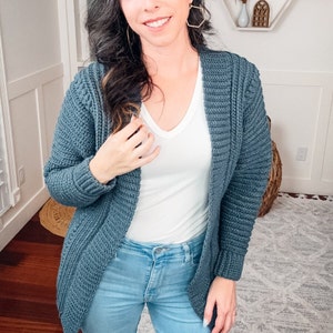 A smiling woman standing indoors wearing a unique jewelry piece, a blue crochet cardigan, white top, and blue jeans.