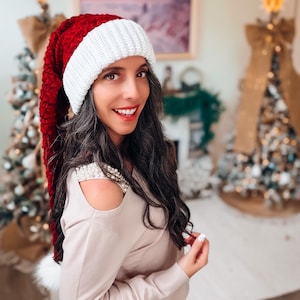 Op de afbeelding: Een lange, dieprode kerstmuts van fluweel met een dikke, wit geribbelde gebreide rand en een witte pompon aan de punt. De muts heeft een zachte, pluche textuur en een klassieke kegelvorm die aan de zijkant naar beneden hangt.