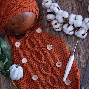 May include: A crocheted baby outfit in a rust orange color with a matching hat. The outfit has a cable knit pattern and is decorated with wooden buttons. A crochet hook is also pictured.