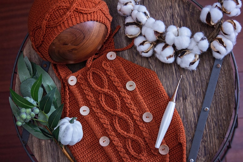 May include: A crocheted baby outfit in a rust orange color with a matching hat. The outfit has a cable knit pattern and is decorated with wooden buttons. A crochet hook is also pictured.