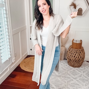 A woman stands in a well-lit room, smiling at the camera, wearing a white top, long beige crochet cardigan, blue jeans, and brown shoes adorned with unique jewellery.