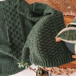 Puede incluir: Una manta de ganchillo de color verde bosque oscuro que presenta un patr&oacute;n de tejido de cesta texturizado en el centro, bordeado por un borde acanalado. La tela muestra una puntada de hilo consistente en toda su superficie y un color s&oacute;lido.