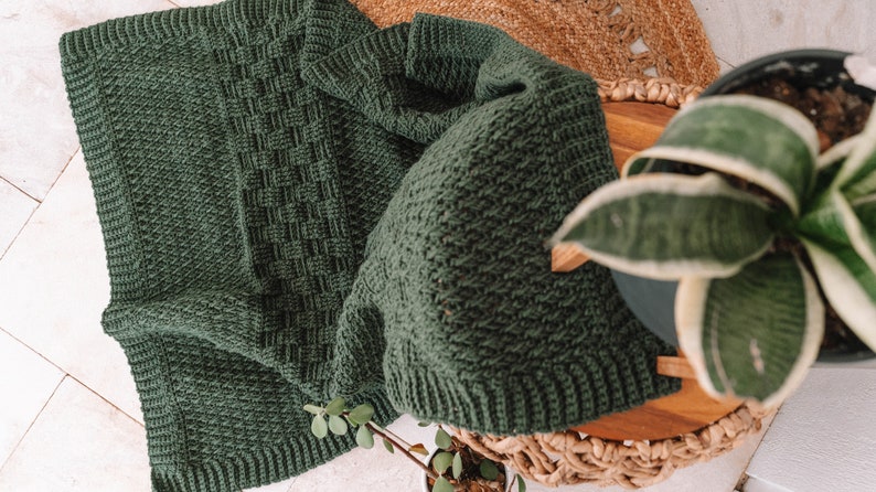 Puede incluir: Una manta de ganchillo de color verde bosque oscuro que presenta un patr&oacute;n de tejido de cesta texturizado en el centro, bordeado por un borde acanalado. La tela muestra una puntada de hilo consistente en toda su superficie y un color s&oacute;lido.