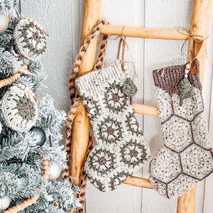 May include: Two handmade crochet Christmas stockings hanging on a wooden ladder. The left stocking features a floral granny square pattern in cream, sage green, and brown. The right stocking has a hexagonal honeycomb pattern with a dark brown cuff.