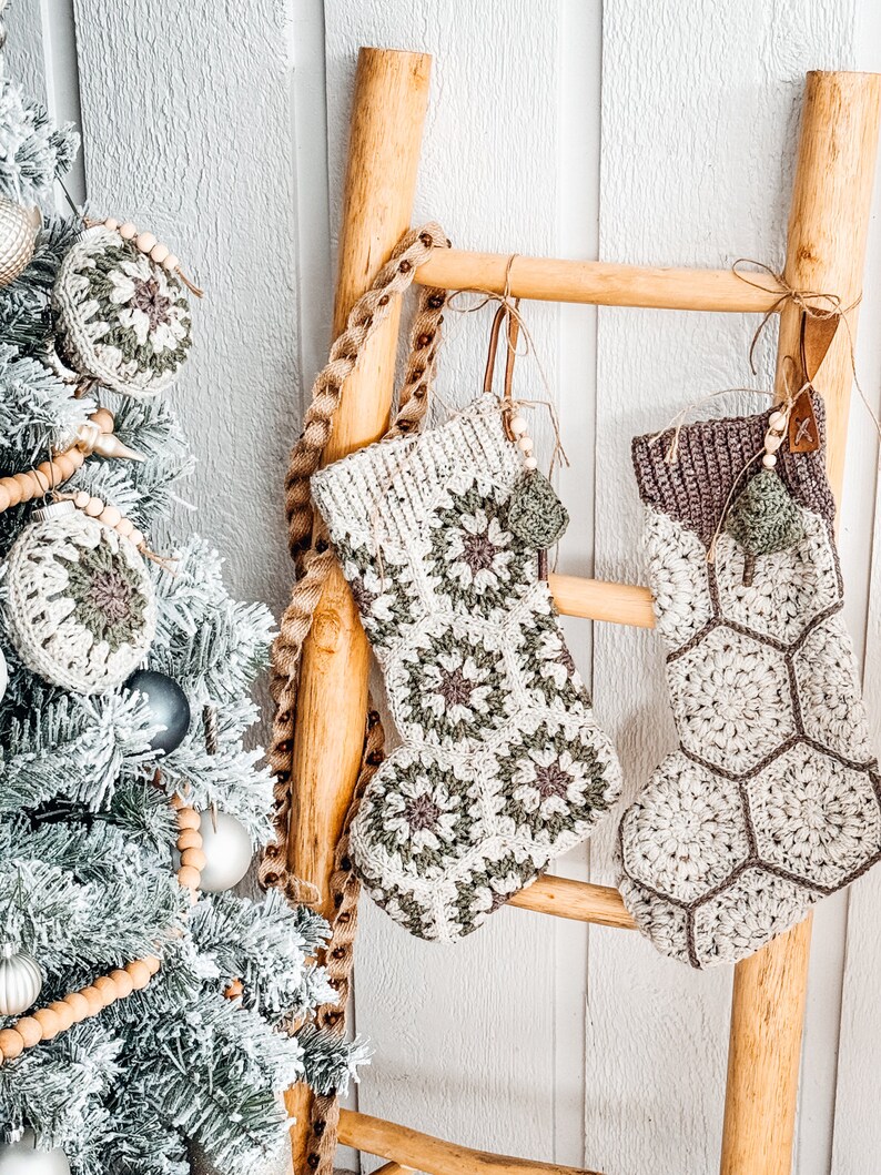 May include: Two handmade crochet Christmas stockings hanging on a wooden ladder. The left stocking features a floral granny square pattern in cream, sage green, and brown. The right stocking has a hexagonal honeycomb pattern with a dark brown cuff.
