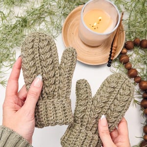 May include: A pair of olive green knitted mittens with a cable knit pattern. The mittens are held by hands against a white background.