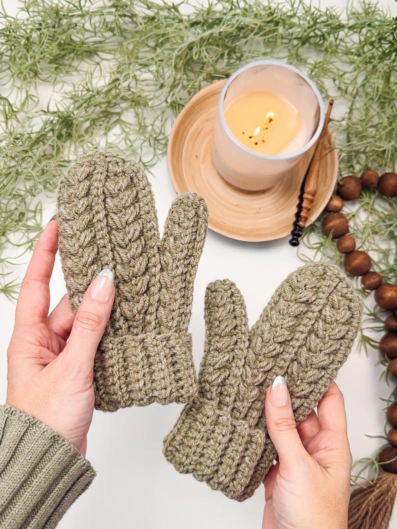May include: A pair of olive green knitted mittens with a cable knit pattern. The mittens are held by hands against a white background.